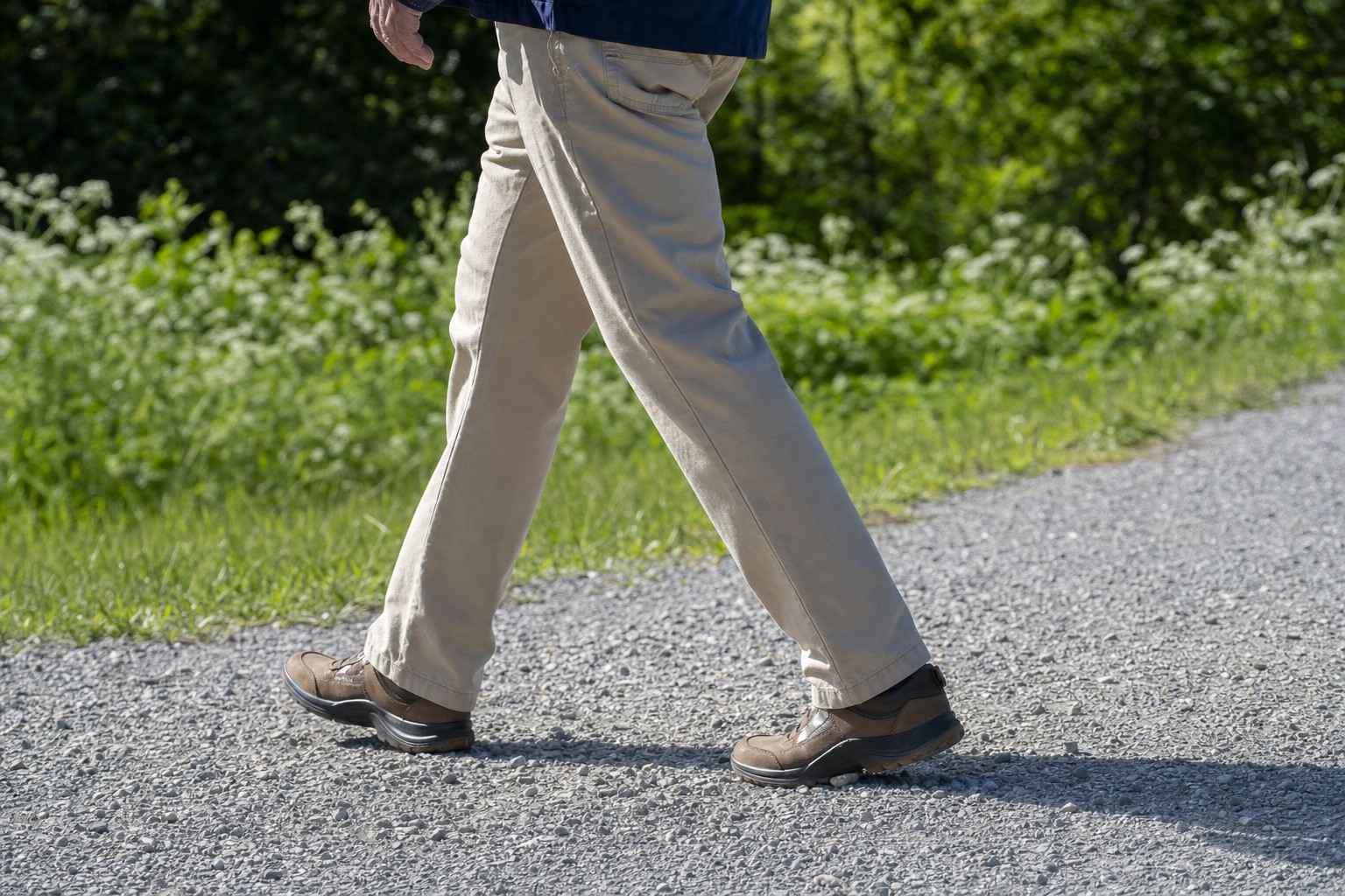 Close side view of an older person's legs mid-stride, walking comfortably on an easy gravel path in bright daylight