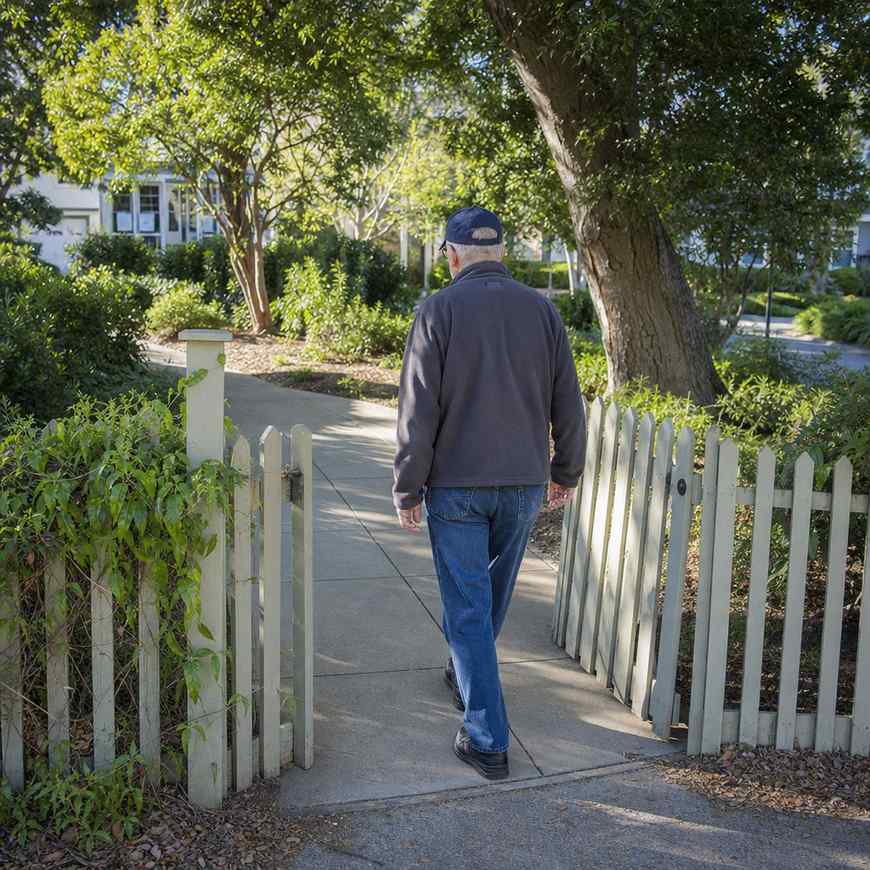 An older man walking a familiar loop past a garden gate, seen from behind in morning light