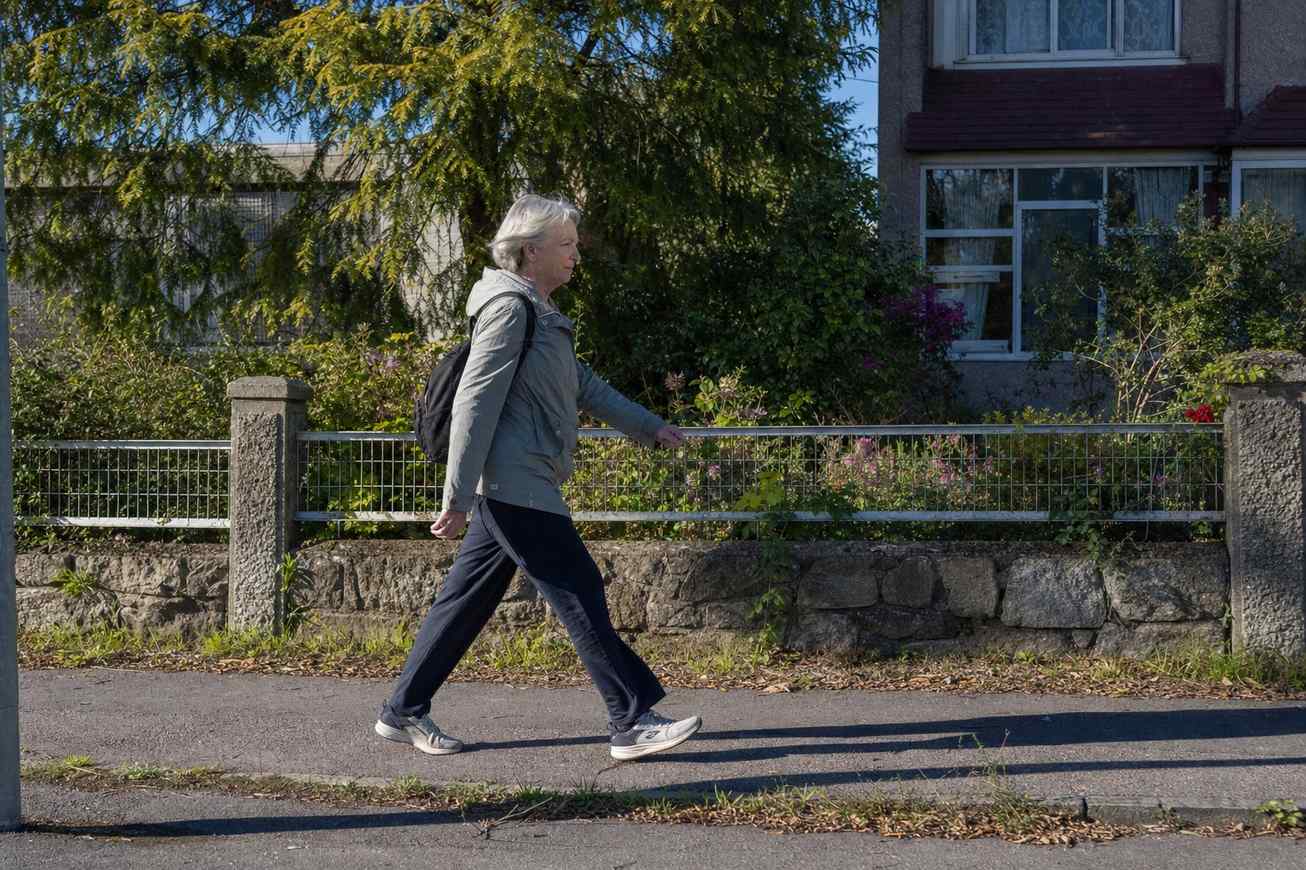 An older woman walking at a brisk purposeful pace along a neighborhood street, seen from the side in bright daylight